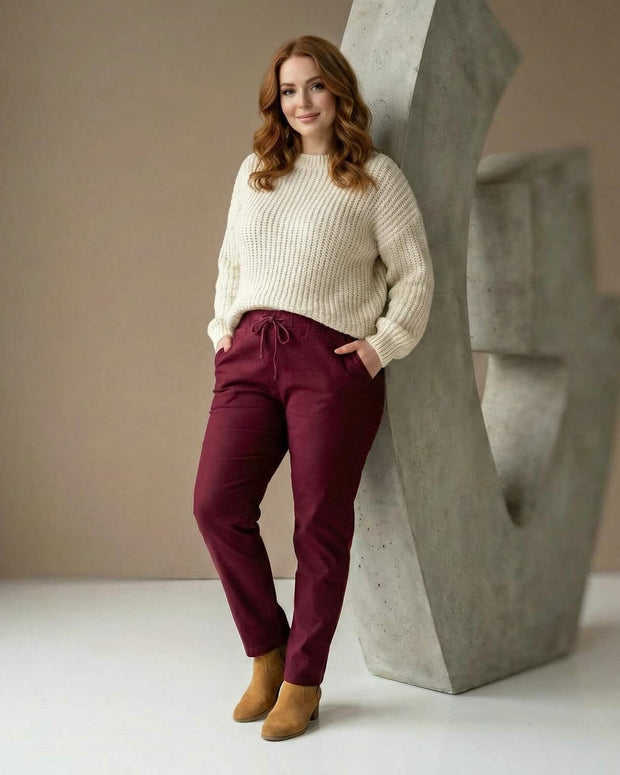 Γυναίκα φοράει μπορντό παντελόνι και λευκό πουλόβερ και στέκεται σε στούντιο με μοντέρνο σκηνικό