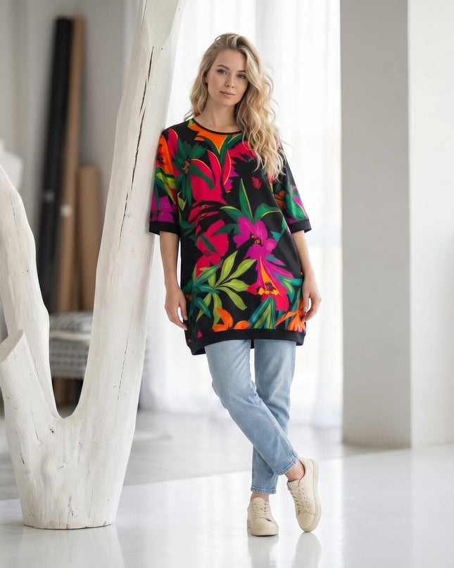 Ολόσωμη λήψη: γυναίκα φοράει πολύχρωμη καλοκαιρινή τουνίκ σε μεγάλα μεγέθη με τροπικό φλοράλ σχέδιο, συνδυασμένη με τζιν παντελόνι και αθλητικά.