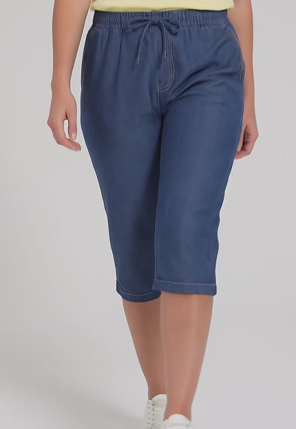 Ένα plus-size μοντέλο σε στούντιο με ουδέτερο φόντο φοράει το Γυναικείο μπλε ελαστικό τζιν παντελόνι 7/8 plus size και περπατάει αργά προς την κάμερα