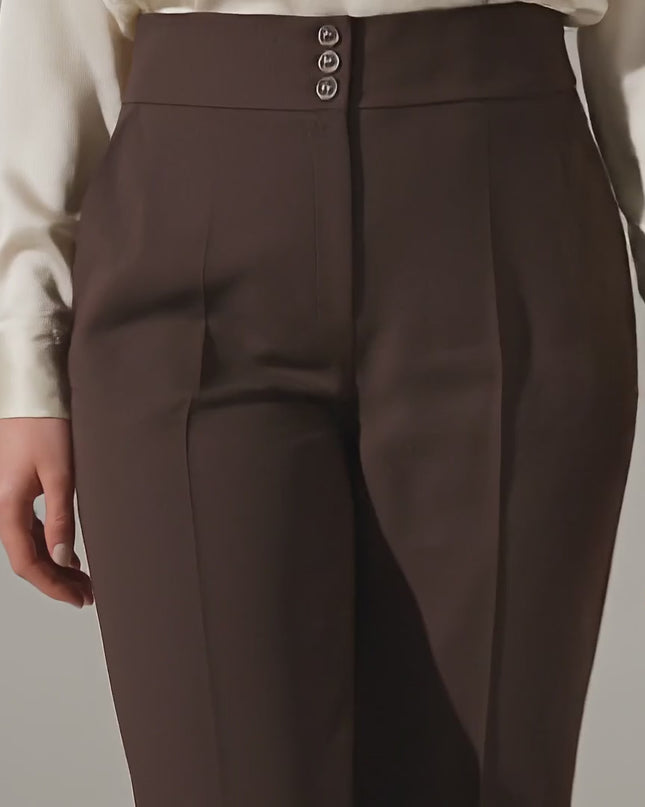 Ένα μοντέλο περπατάει αργά προς την κάμερα σε περιβάλλον στούντιο φορώντας το Κομψό καφέ ψηλόμεσο παντελόνι με κουμπιά σε μεγάλα μεγέθη