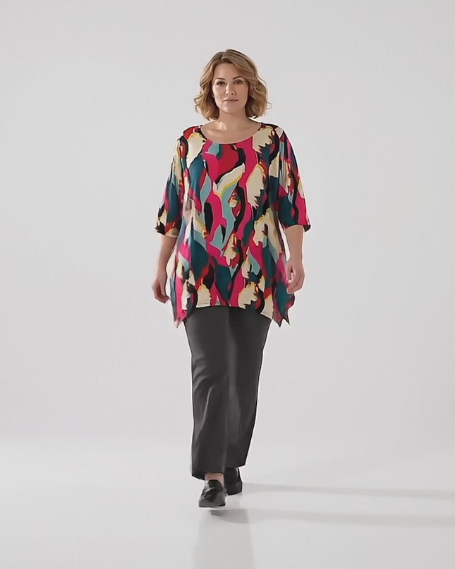 Σε ένα περιβάλλον στούντιο με ουδέτερο φόντο ένα plus size μοντέλο περπατάει αργά προς την κάμερα φορώντας το προϊόν Μακριά καλοκαιρινή plus size τουνίκ με πολύχρωμο αφηρημένο μοτίβο