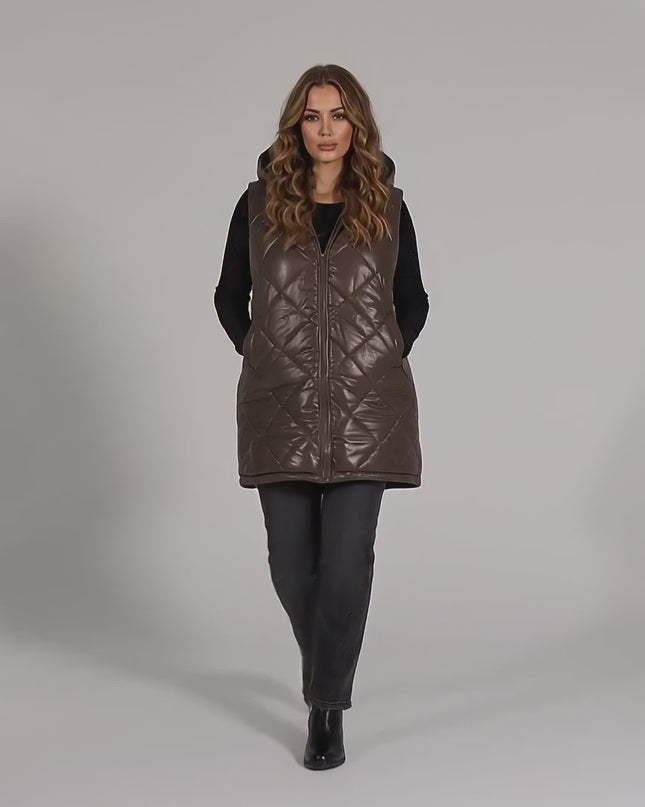 Βίντεο παρουσίασης σε περιβάλλον στούντιο με ουδέτερο φόντο όπου ένα plus-size μοντέλο περπατάει αργά προς την κάμερα φορώντας το Μακρύ καπιτονέ γιλέκο με κουκούλα σε σοκολατί απόχρωση