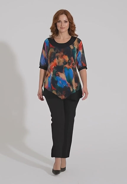 Ένα plus size μοντέλο περπατάει αργά προς την κάμερα σε περιβάλλον στούντιο με ουδέτερο φόντο φορώντας το προϊόν Επίσημη μαύρη τουνίκ με ασύμμετρο σιφόν και πολύχρωμο print