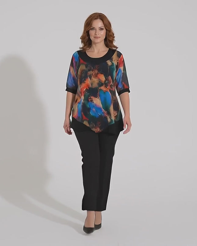 Ένα plus size μοντέλο περπατάει αργά προς την κάμερα σε περιβάλλον στούντιο με ουδέτερο φόντο φορώντας το προϊόν Επίσημη μαύρη τουνίκ με ασύμμετρο σιφόν και πολύχρωμο print