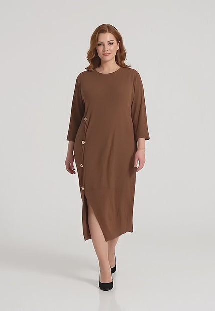 Ένα plus-size μοντέλο περπατάει αργά προς την κάμερα σε περιβάλλον στούντιο με ουδέτερο φόντο φορώντας το προϊόν Επίσημο plus size μάξι φόρεμα καφέ με διακοσμητικά κουμπιά