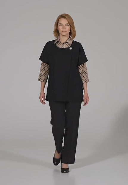 Ένα plus-size μοντέλο περπατάει αργά προς την κάμερα σε περιβάλλον στούντιο με ουδέτερο φόντο φορώντας το Επίσημο σετ μεγάλα μεγέθη με μπεζ πουά πουκάμισο και μαύρο γιλέκο