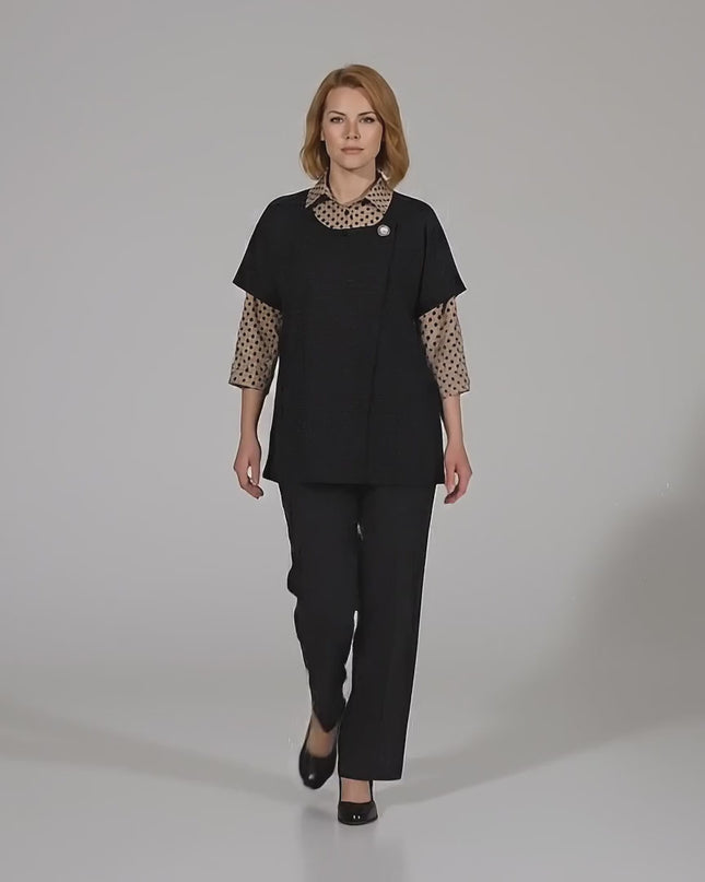 Ένα plus-size μοντέλο περπατάει αργά προς την κάμερα σε περιβάλλον στούντιο με ουδέτερο φόντο φορώντας το Επίσημο σετ μεγάλα μεγέθη με μπεζ πουά πουκάμισο και μαύρο γιλέκο