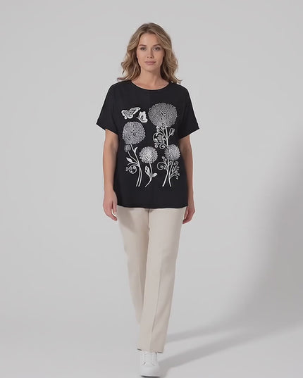 Ένα plus-size μοντέλο περπατάει αργά προς την κάμερα σε περιβάλλον στούντιο με ουδέτερο φόντο, φορώντας το προϊόν Μαύρη καλοκαιρινή μάξι τουνίκ με εντυπωσιακό απλικέ λουλουδιών