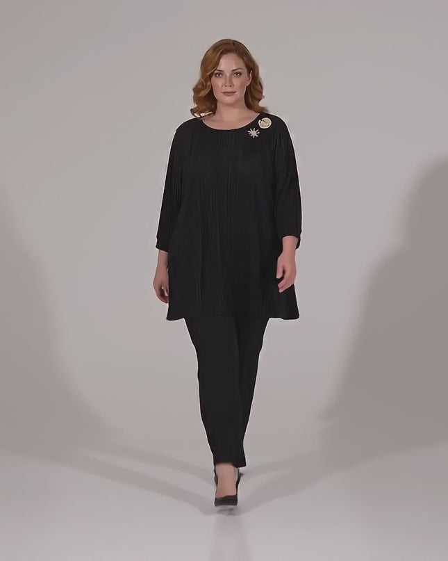 Βίντεο παρουσίασης σε στούντιο με ουδέτερο φόντο όπου ένα μοντέλο περπατάει αργά προς την κάμερα φορώντας Επίσημη μαύρη τουνίκ σε μεγάλα μεγέθη από πλισέ σολεΐ με καρφίτσα