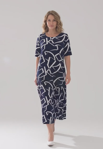 Ένα plus size μοντέλο περπατάει αργά προς την κάμερα σε περιβάλλον στούντιο φορώντας το Σκούρο μπλε επίσημο μάξι φόρεμα με γεωμετρικό print