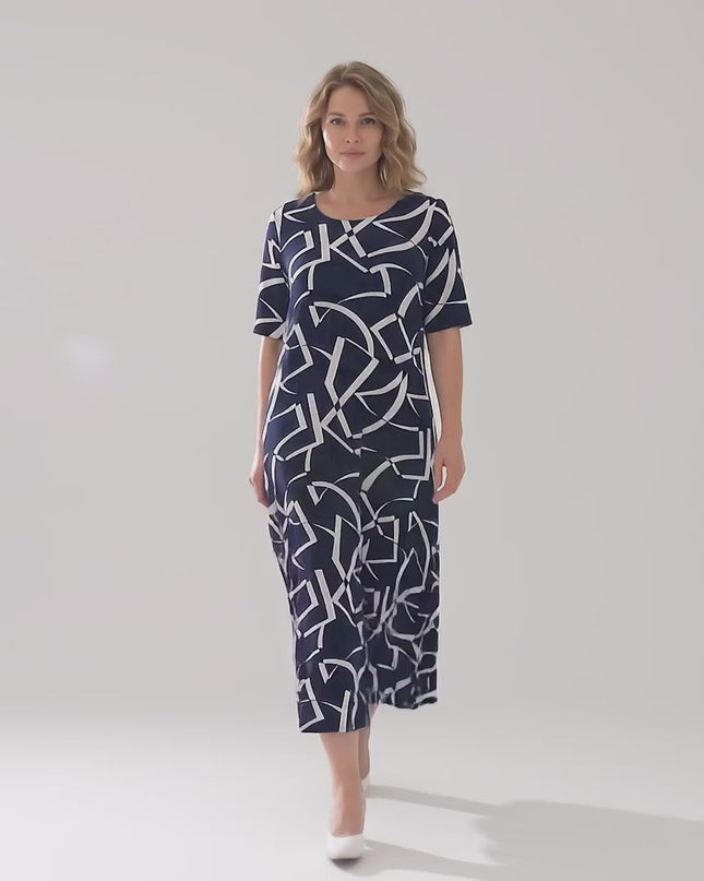 Ένα plus size μοντέλο περπατάει αργά προς την κάμερα σε περιβάλλον στούντιο φορώντας το Σκούρο μπλε επίσημο μάξι φόρεμα με γεωμετρικό print