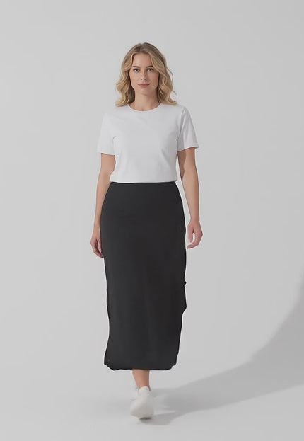 Ένα plus-size μοντέλο φοράει το προϊόν Μαύρη εβαζέ μάξι φούστα μεγάλα μεγέθη με ασύμμετρη γραμμή και περπατάει αργά προς την κάμερα σε ένα περιβάλλον στούντιο με ουδέτερο φόντο