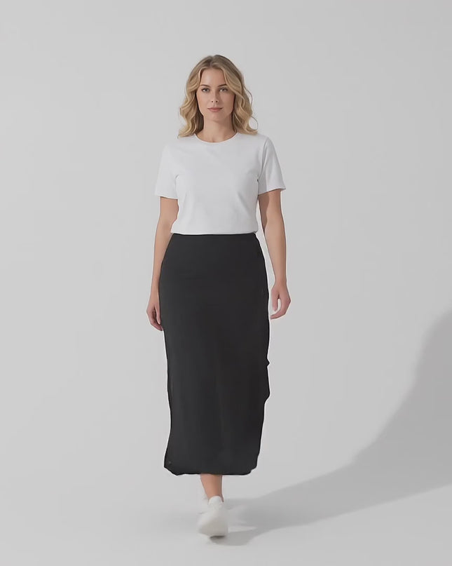 Ένα plus-size μοντέλο φοράει το προϊόν Μαύρη εβαζέ μάξι φούστα μεγάλα μεγέθη με ασύμμετρη γραμμή και περπατάει αργά προς την κάμερα σε ένα περιβάλλον στούντιο με ουδέτερο φόντο