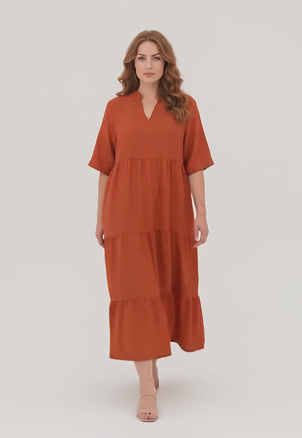 Ένα plus-size γυναικείο μοντέλο σε περιβάλλον στούντιο με ουδέτερο φόντο περπατάει αργά προς την κάμερα φορώντας το Επίσημο Μάξι Φόρεμα Κεραμιδί σε Μεγάλα Μεγέθη