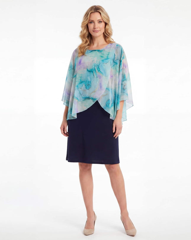 Ολόσωμη λήψη γυναίκας που φοράει σκούρο μπλε επίσημο φόρεμα σε μεγάλα μεγέθη, με αέρινη ασύμμετρη κάπα από σιφόν με αφηρημένο print σε λευκό φόντο.