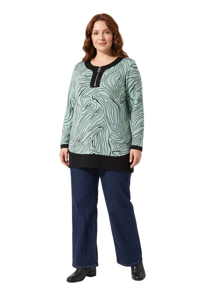 Γυναίκα φορά ανοιχτοπράσινη τουνίκ με μαύρες αφηρημένες γραμμές, μαύρο τελείωμα και κοντό μπροστινό φερμουάρ, με μπλε τζιν και μαύρα μποτάκια.